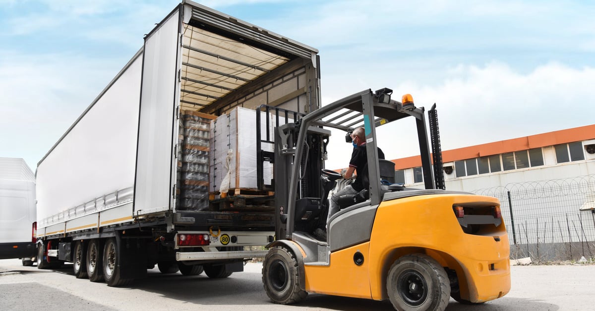 A yellow forklift lifts a pallet of white boxes wrapped in saran wrap into a partially filled semi truck trailer.