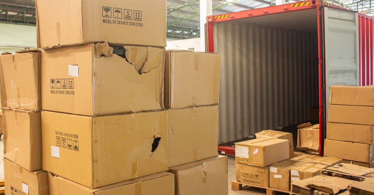 unloading carton from container and carton damage from loading or transport process. Stacks of unloaded, damaged cardboard boxes near an open red shipping container with cardboard boxes inside.