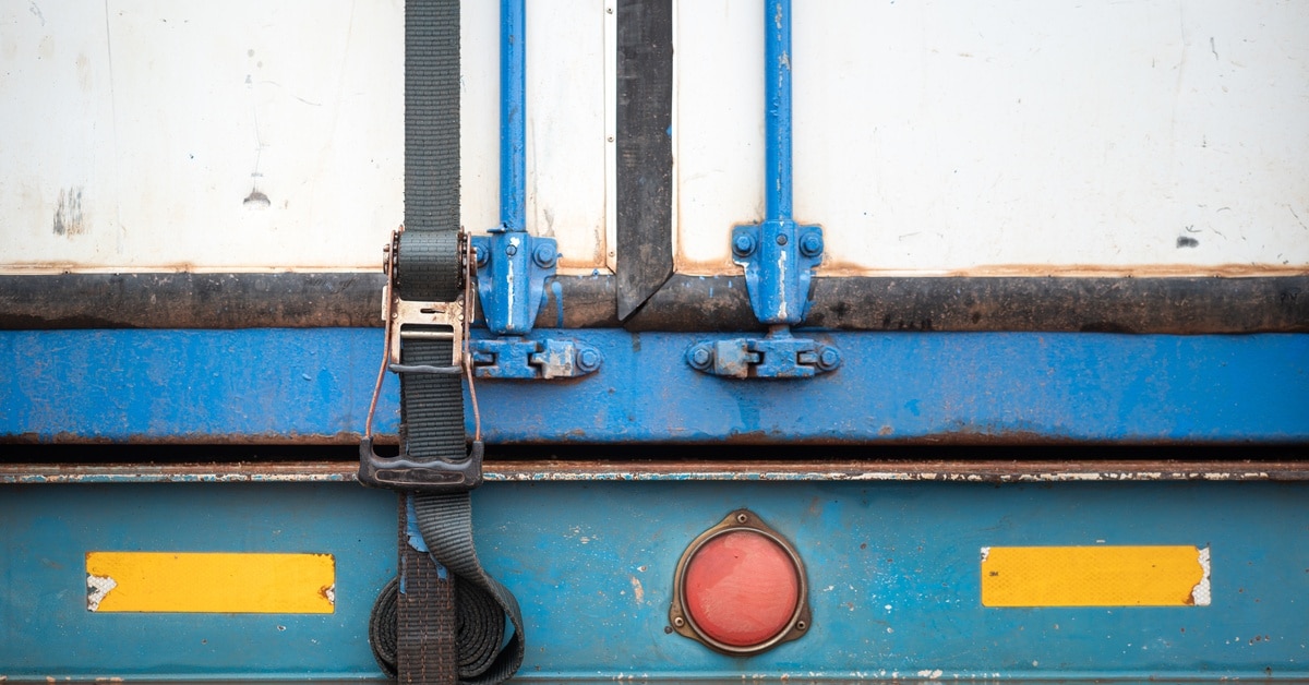 4 Environmental Benefits of Securing Intermodal Containers The back of a white cargo shipment trailer with a webbing belt strap that is latched down to secure the door.