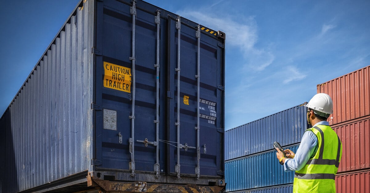 A man wearing a bright vest and hardhat stands behind a large shipping truck while holding a cellphone.