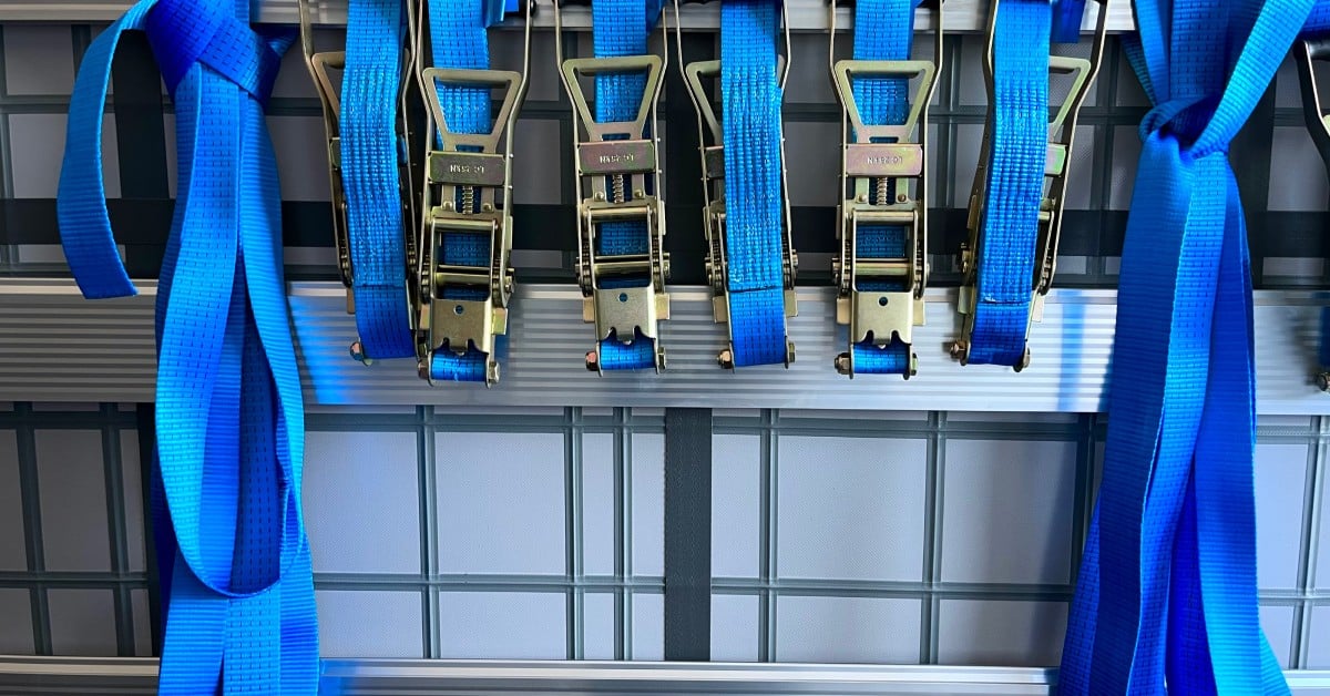 LogistickInc-372788-blue-ratchet-straps-image1 A group of multiple blue-colored ratchet tie down straps hanging next to one another. The straps have silver buckles.