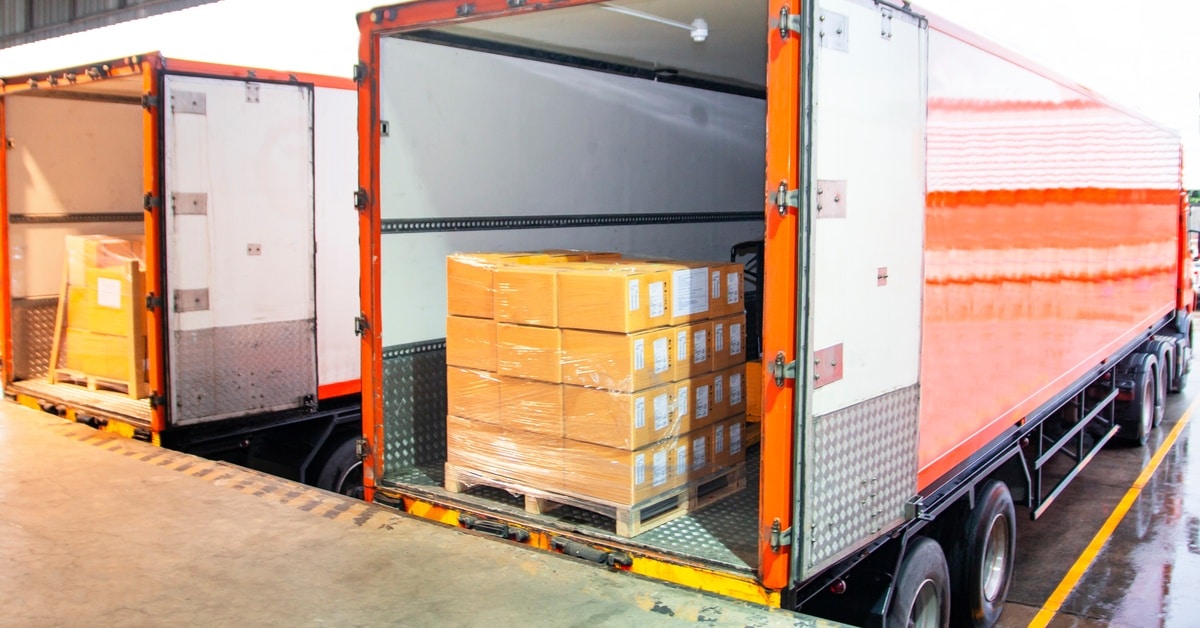 Understanding the Need for Adequate Load Securement Two orange shipping trucks filled with pallets of cardboard boxes are parked at a warehouse loading dock.