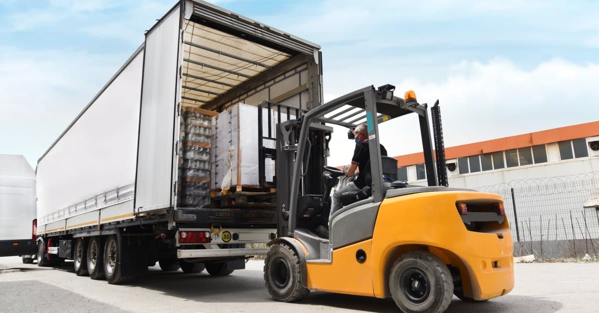 When To Replace Your Cargo Load Bars: Signs of Wear and Tear A yellow forklift is parked next to a large shipping truck while loading pallets of cargo. Load bars hold the cargo.