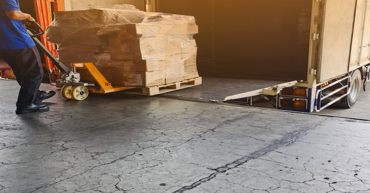A worker wearing a blue shirt uses a dolly to load multiple pallets of cargo onto a truck parked at a loading dock.